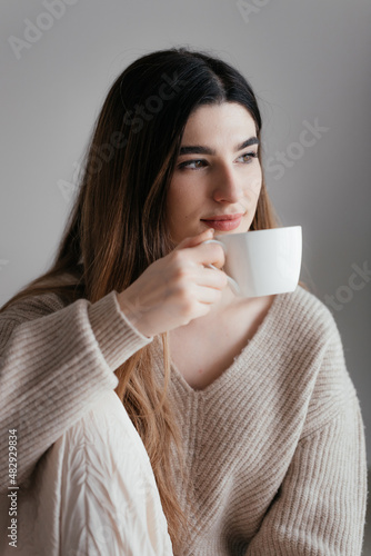 Beautiful young girl with brown hair in beige sweater looks away and holds a white cup in her hand. Portrait of girl drinking coffee at home. Woman in warm sweater with cup of hot drink looking away