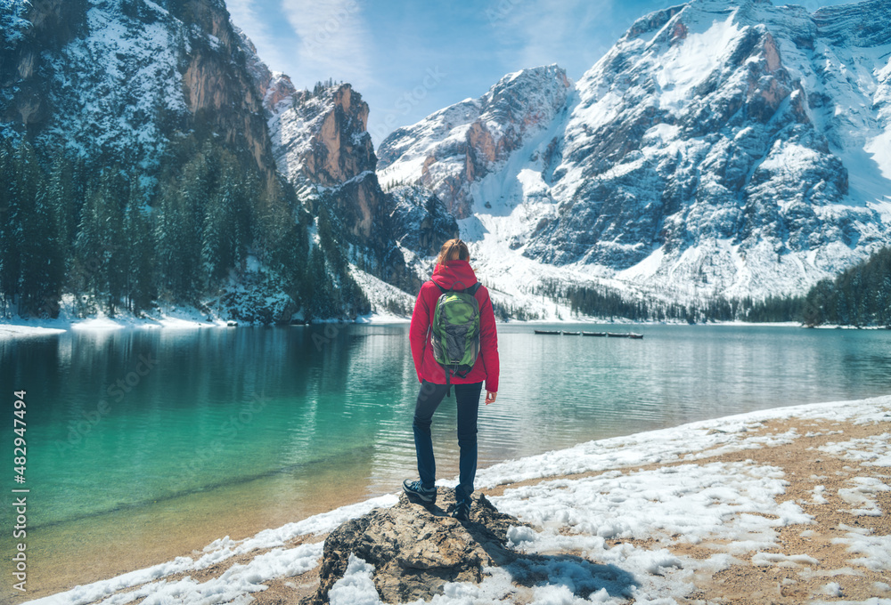 Young woman with backpack on the stone and snowy shore of Braies lake with clear water at sunset in spring. Landscape with girl, reflection in water, rocks in snow,  mountains, green trees, blue sky