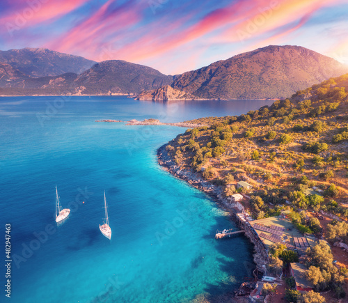 Fototapeta Naklejka Na Ścianę i Meble -  Aerial view of beautiful yachts and boats on the sea at sunset in summer in Turkey. Top view of luxury yacht, sailboat, clear blue water, rocks, colorful sky, mountain, green trees. Travel. Landscape
