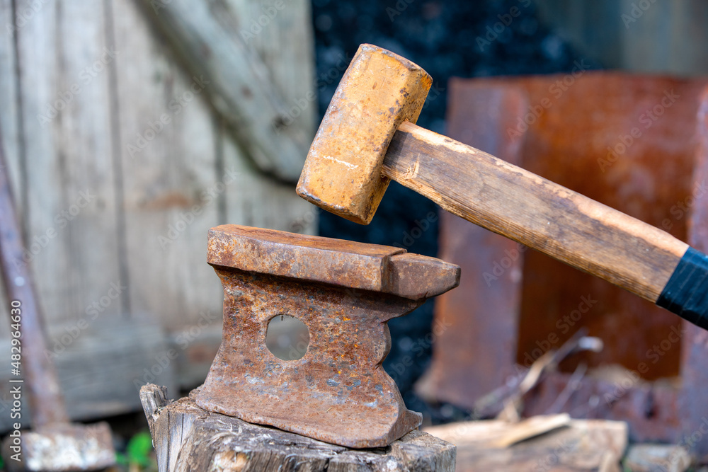 Old rusty anvil stands on a stump in the backyard of village house, sledgehammer leaning on wooden fence enclosing a barn with coal on blurred background. Heavy working tools Old rusty anvil stands on a stump in the backyard of village house, sledgehammer leaning on wooden fence enclosing a barn with coal on blurred background. Heavy working tools