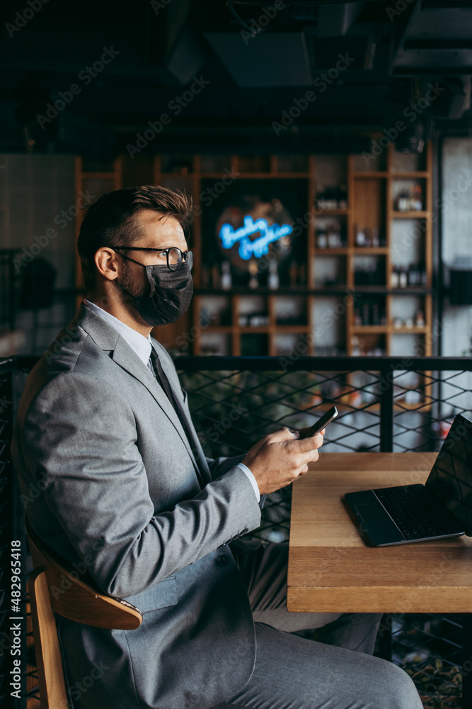 A businessman with a protective mask on his face is sitting in a cafe and talking on the phone. Coronavirus, Covid-19 concept.