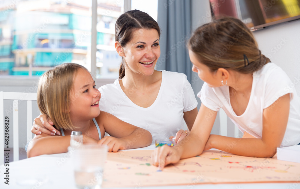 Mom with two daughters play board game at the table at home