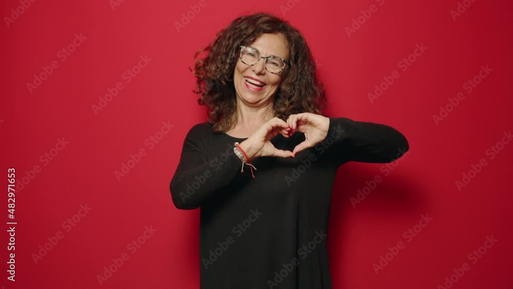 Middle age woman smiling confident doing heart symbol with hands over red background Middle age woman smiling confident doing heart symbol with hands over red background