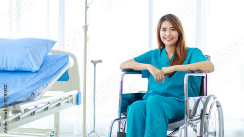 Portrait shot of young happy healthy Asian female patient in blue green hospital uniform sitting alone smiling look at camera on wheelchair in wardroom with bed and clinical saline solution stand photo