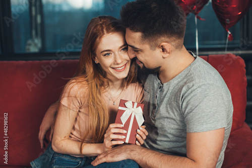 Caucasian couple, girl holding in hands red gift box for anniversary congrats sitting on sofa. Happy birthday. Celebrate party. Valentines day. Helium balloons.