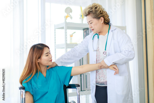 Old senior professional female doctor in white lab coat standing using stethoscope listening to heartbeat of disabled handicapped woman patient sitting on wheelchair in hospital checkup ward room photo