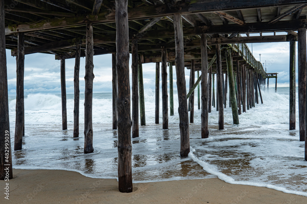 walk under the creaking wooden fisherman's pier on the beach, with foam washed in with each crashing wave on the wet sand, soaking the planks of wood and depositing salty water while eroding the shore