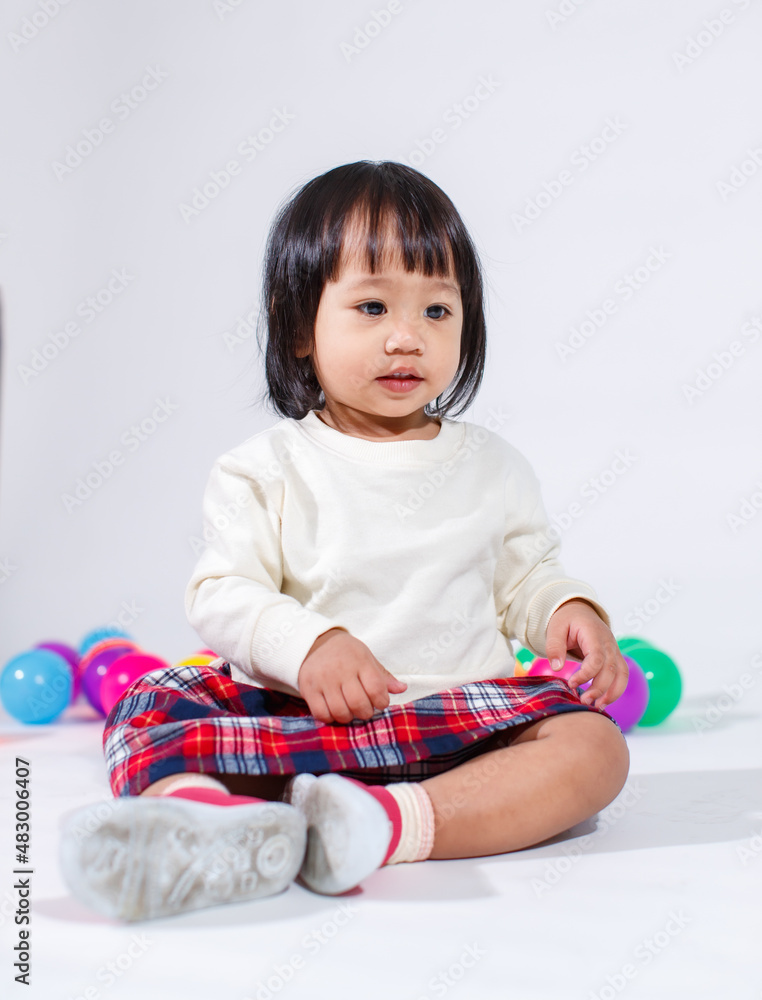 Naklejka premium Studio shot of little cute short black hair Asian baby girl daughter model in casual plaid skirt sitting on floor smiling laughing playing with colorful round balls toy alone on white background