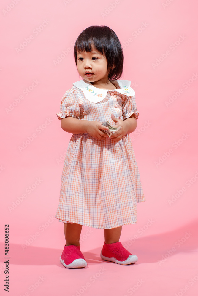 Portrait studio shot little cute Asian kindergarten preschooler girl daughter model in casual long dress and shoes standing look at camera holding hundred dollars banknote in hands on pink background Portrait studio shot little cute Asian kindergarten preschooler girl daughter model in casual long dress and shoes standing look at camera holding hundred dollars banknote in hands on pink background