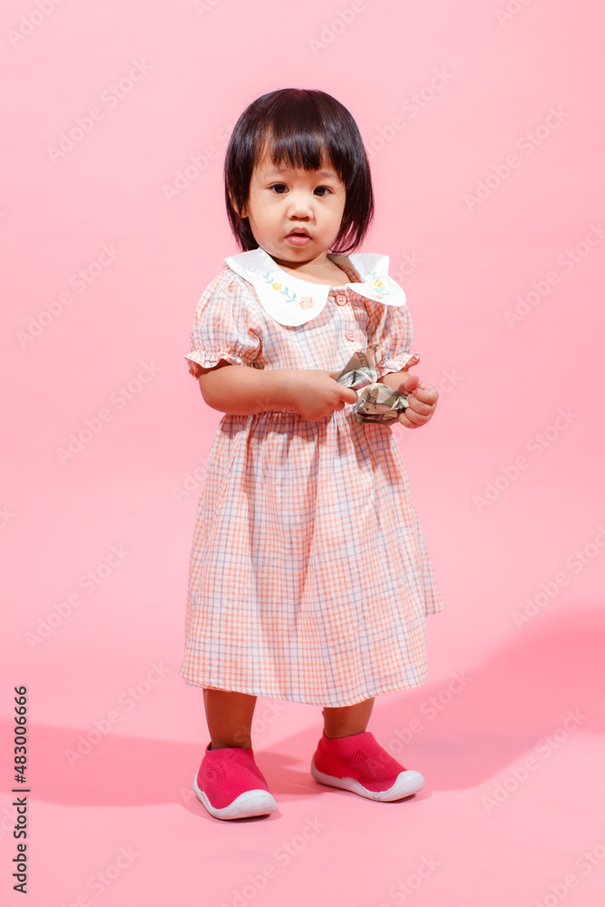Portrait studio shot little cute Asian kindergarten preschooler girl daughter model in casual long dress and shoes standing look at camera holding hundred dollars banknote in hands on pink background Portrait studio shot little cute Asian kindergarten preschooler girl daughter model in casual long dress and shoes standing look at camera holding hundred dollars banknote in hands on pink background