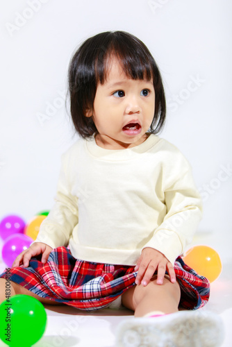 Studio shot of little cute short black hair Asian baby girl daughter model in casual plaid skirt sitting on floor smiling laughing playing with colorful round balls toy alone on white background