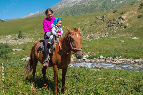 Mother with child are sitting on horse back with green mountains background. © Adil