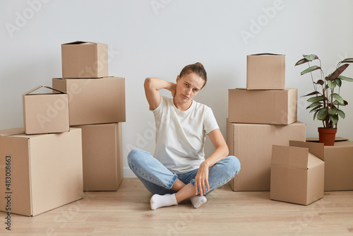 Portrait if sick tired woman wearing white T-shirt sitting on the floor near cardboard boxes with personal pile, being tired during moving in to new house, feeling pain in neck.