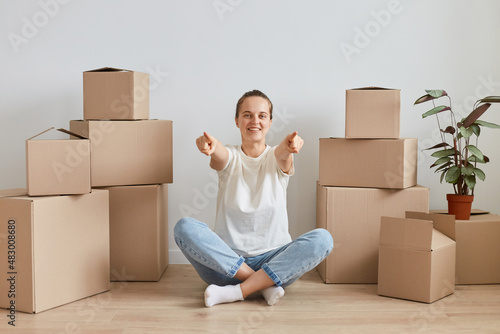 Smiling happy young adult woman wearing white T-shirt sitting on the floor near cardboard boxes with stuff, posing in lotus pose and pointing fingers to camera, choosing you.