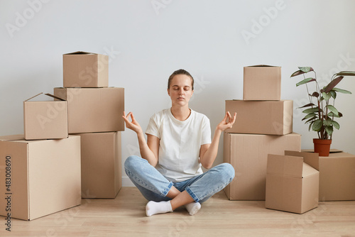 Indoor shot of tired exhausted woman wearing white T-shirt sitting on the floor near cardboard boxes with belongings, trying to relax with yoga practice during relocating to new flat.