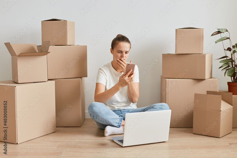 Sad upset young adult Caucasian woman wearing white T-shirt sitting on the floor near cardboard boxes, using mobile phone, looking at display and expressing sorrow. Sad upset young adult Caucasian woman wearing white T-shirt sitting on the floor near cardboard boxes, using mobile phone, looking at display and expressing sorrow.