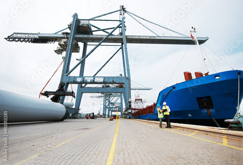 That makes you larger than life. An anchored ship on the dock with cargo being loaded on to it with cranes and two people standing next to the ship.