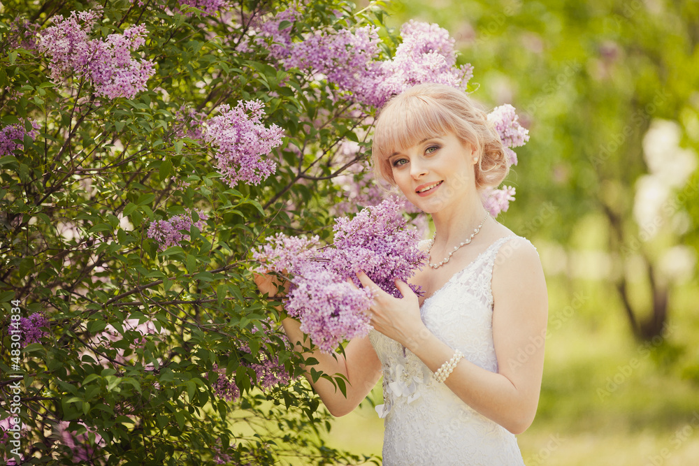 Fototapeta premium Beautiful blonde Bride in elegant wedding dress in blooming lilac garden. Waist portrait in sunset light. Pretty young caucasian girl. Spring wedding day. Romantic mood. Romantic wedding ceremony