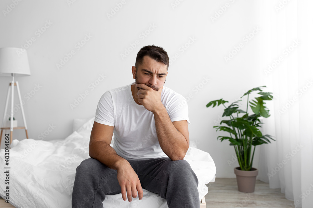 Unhappy sad millennial european muscular male sits on white bed and suffers from stress and thinks Unhappy sad millennial european muscular male sits on white bed and suffers from stress and thinks