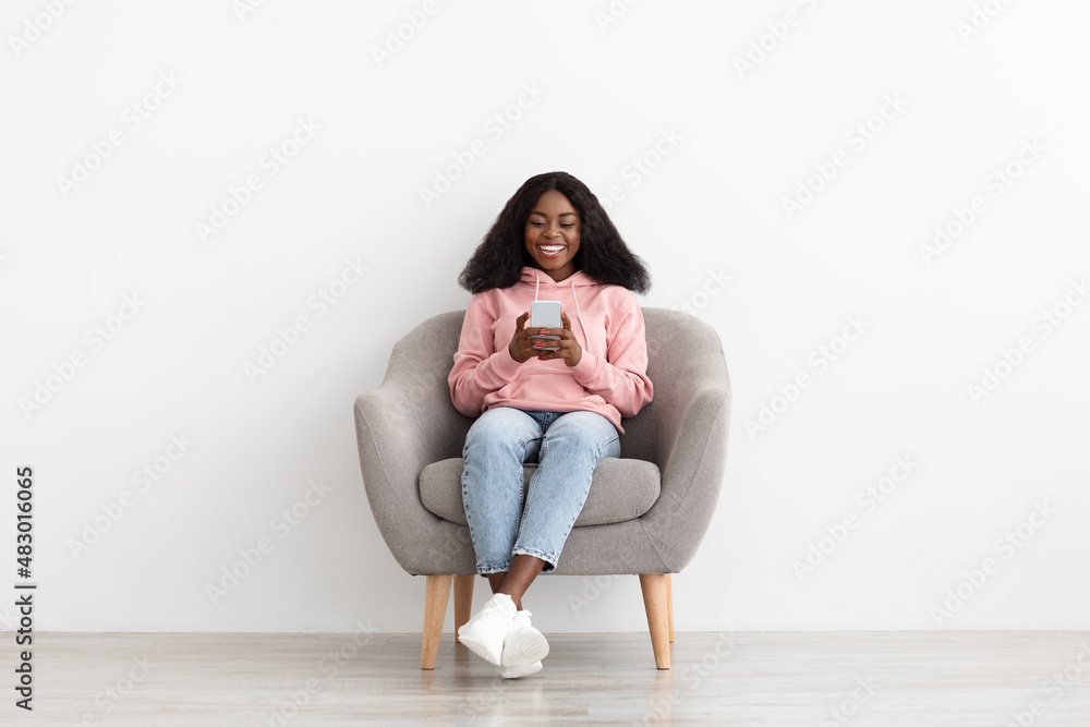 Cheerful black woman chilling at arm chair with smartphone Cheerful black woman chilling at arm chair with smartphone