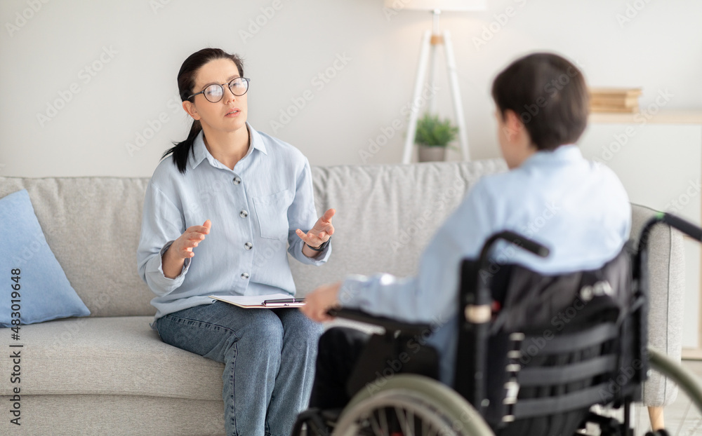 Psychotherapy for disabled teens. Young female psychologist speaking with teenage boy in wheelchair at office Psychotherapy for disabled teens. Young female psychologist speaking with teenage boy in wheelchair at office