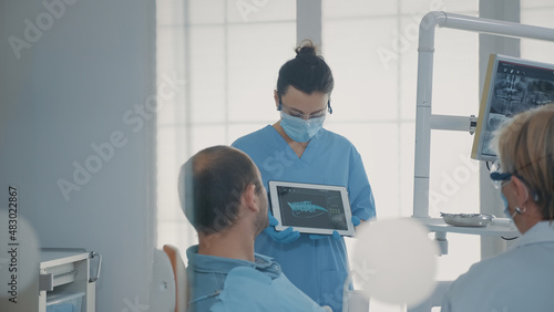 Orthodontic nurse holding x ray results on tablet to show and explain diagnosis to patient in dentistry office. Team of experts showing teeth radiography on device  talking about implant.