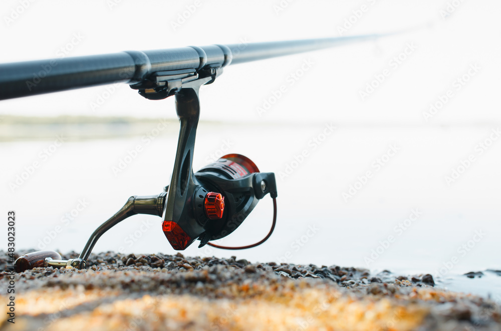 Fishing hobby and sport concept. Spinning rod, reel on shore near water outdoors, close-up. Selective soft focus Fishing hobby and sport concept. Spinning rod, reel on shore near water outdoors, close-up. Selective soft focus