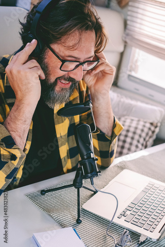 Modern adult man in podcast recording activity sitting at the table. Online job using laptop computer and connection technology. Freelance at work indoor in alternative office photo