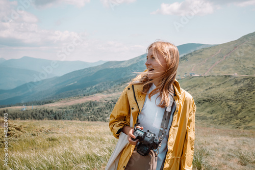 Beautiful smile blonde girl tourist in yellow raincoat and with a camera looks at the beautiful green hills of the Carpathians of Ukraine on a sunny day
