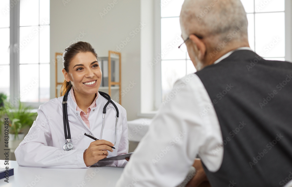 Friendly young female doctor in hospital office consults senior male patient and makes entries in his medical card. Smiling nurse sitting at table talking to mature man sitting with his back to camera Friendly young female doctor in hospital office consults senior male patient and makes entries in his medical card. Smiling nurse sitting at table talking to mature man sitting with his back to camera