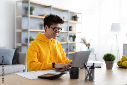 Smiling asian man in glasses working on laptop at home