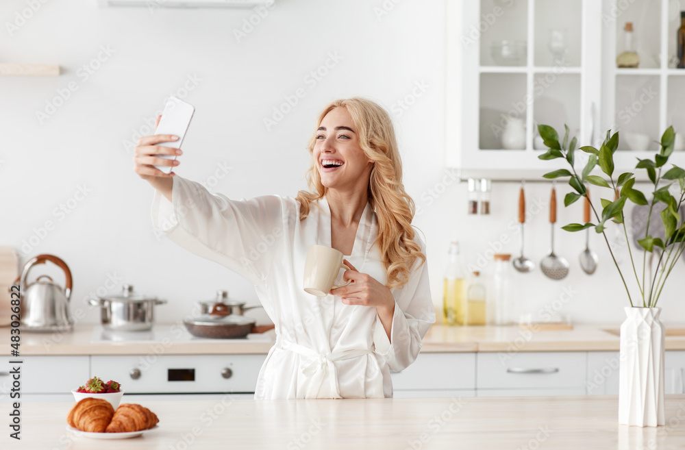 Smiling pretty young european blonde female in robe drink coffee, taking selfie on smartphone at breakfast Smiling pretty young european blonde female in robe drink coffee, taking selfie on smartphone at breakfast