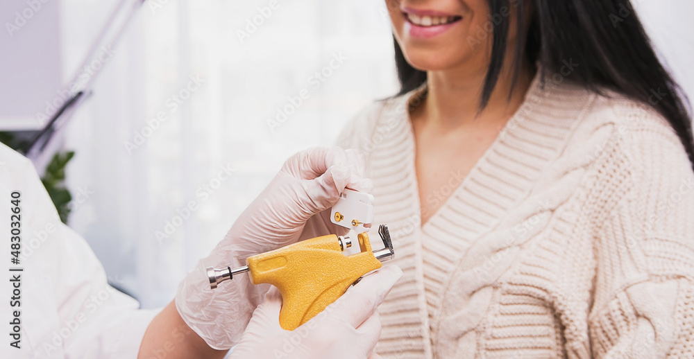 Specialist in medical gloves holding in her hands piercing gun and medical earrings showing to a woman client in the cosmetologist cabinet. Specialist in medical gloves holding in her hands piercing gun and medical earrings showing to a woman client in the cosmetologist cabinet.