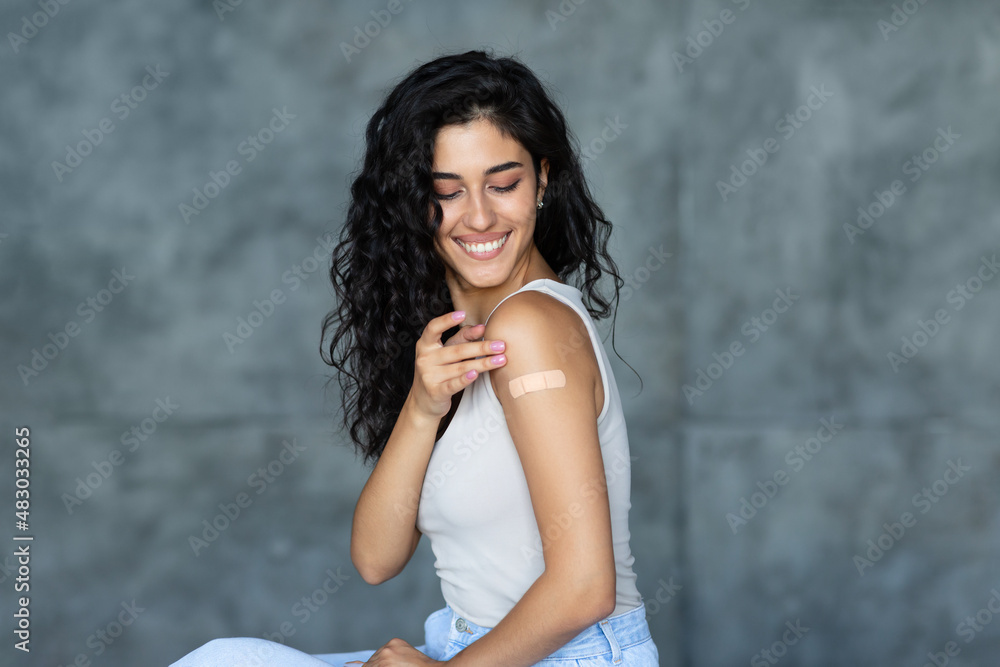 Fototapeta premium Young Caucasian woman showing adhesive bandage on her arm, getting vaccinated against covid-19 on grey background