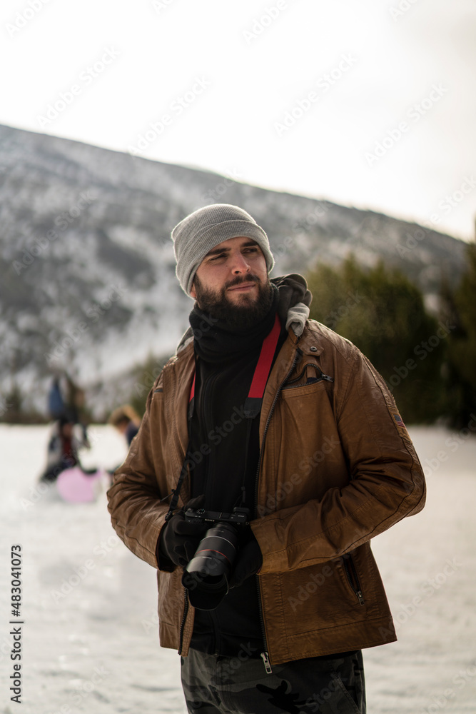 Chico con barba en la nieve con camara de fotos con ropa marron