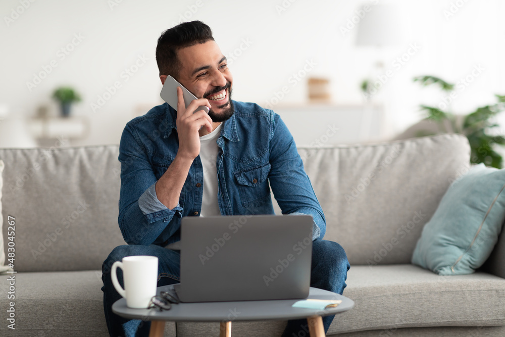 Handsome young Arab guy talking on cellphone while working with laptop at home office, copy space Handsome young Arab guy talking on cellphone while working with laptop at home office, copy space