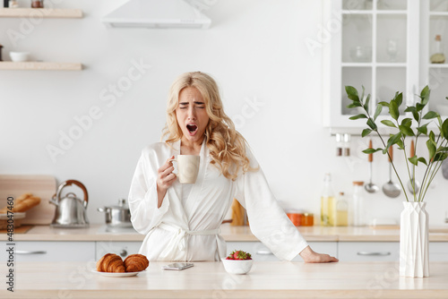 Sleepy tired lazy young pretty caucasian blonde lady in bathrobe wakes up early, drink coffee and yawns