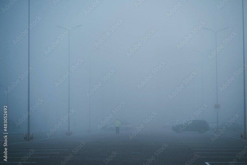 A rear view of parking security man walking over parking area on a foggy autumn morning. Security guard is protecting property from illegal parking