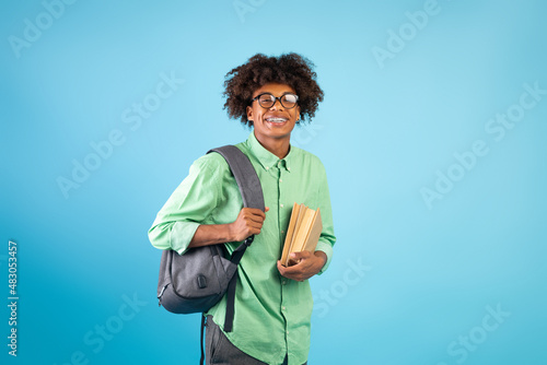 Happy black guy student with backpack and books smiling at camera over blue studio background