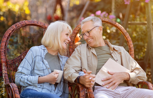 Romantic senior couple sitting in garden in wicker chairs, looking at eah other with love and enjoying warm autumn days