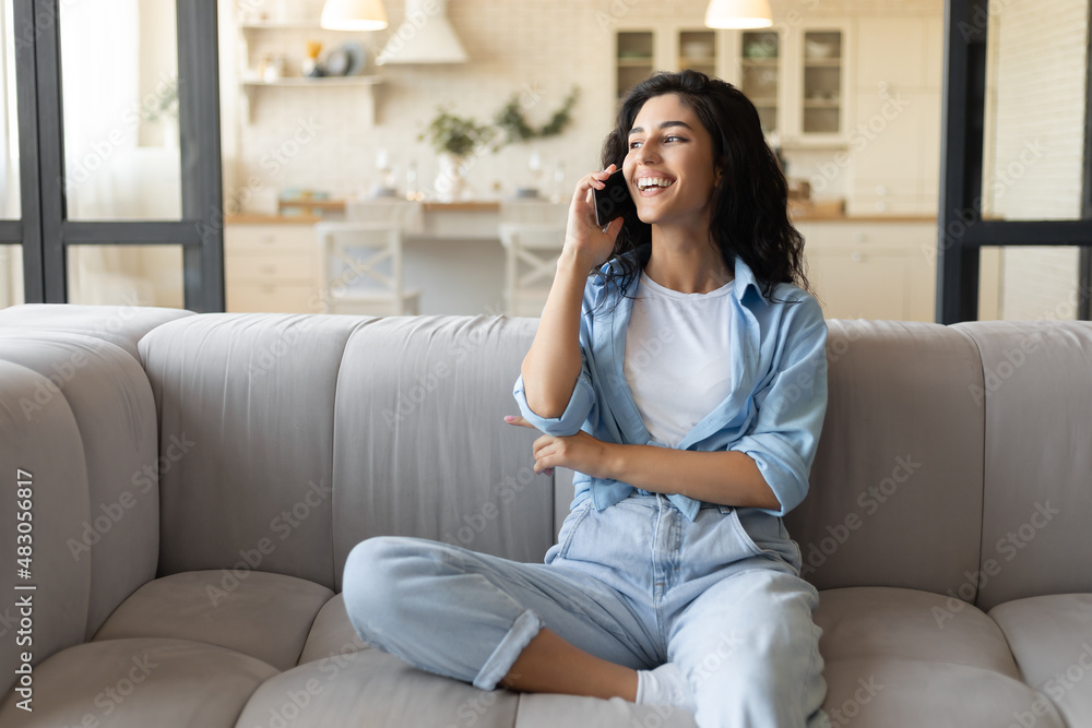 Fototapeta premium Happy young Caucasian lady sitting on couch, having mobile phone conversation at home, free space