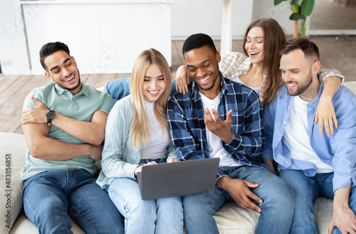 Cheerful diverse friends using laptop pc, sitting on sofa in living room, having video call, chatting online at home