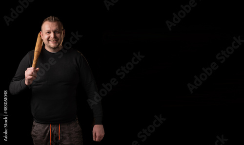 A young man holds a bat in his hand and smiles. Dark background. Place for text. Man in black clothes. Bouncer, security guard, bodyguard concept. photo