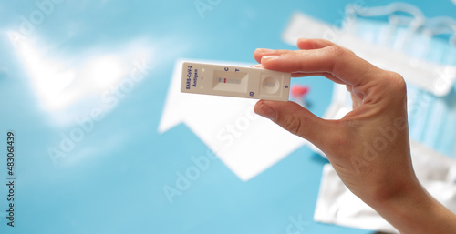 Close up hand of woman holds antigen test on coronavirus. Make fast test at home and get results. On pale blue blurred background lies medical masks and other packages with tests.