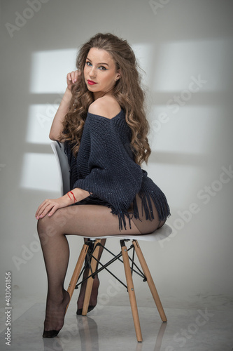 Attractive woman in black pantyhose and long legs posing challenging in front of a background. Classic boudoir scene. Indoor erotic photo. Sensual young  woman with long curly hair sit on the chair