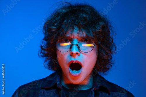 Shocked young man, student with open mouth looking at camera isolated on blue background in neon. Concept of emotions, study, education, fashion