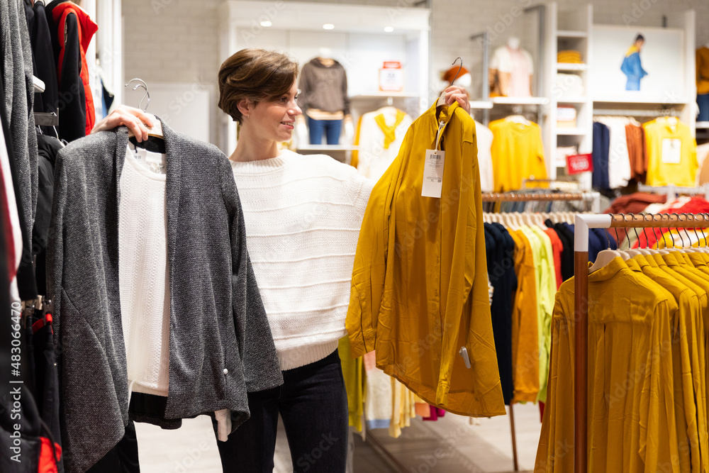 Photo of young attractive brunette woman with a short haircut in a white sweater chooses stylish and casual clothes in a store in a shopping mall. Shop concept