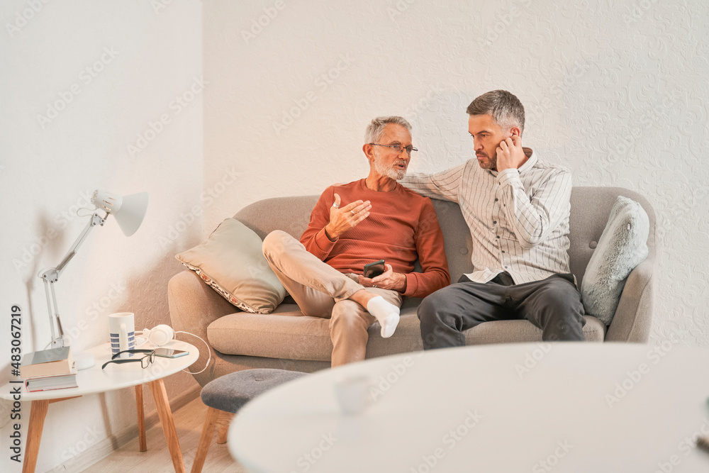 Obraz premium Retirement man with his adult son relaxing on sofa at their cozy apartments
