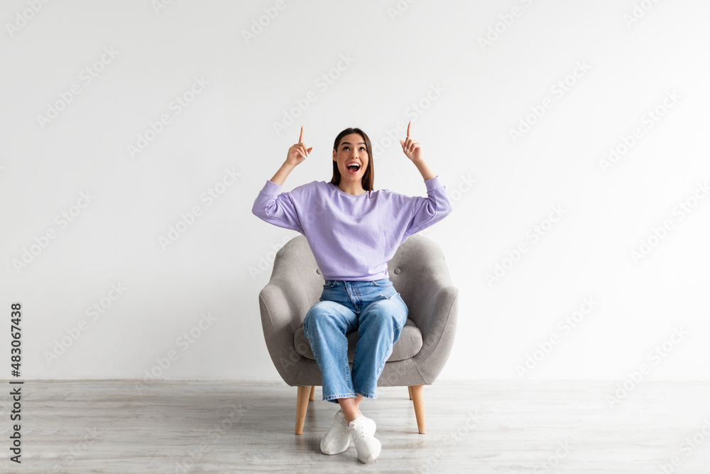 Excited Caucasian woman having idea and pointing two fingers up at copy space, sitting in armchair, mockup Excited Caucasian woman having idea and pointing two fingers up at copy space, sitting in armchair, mockup