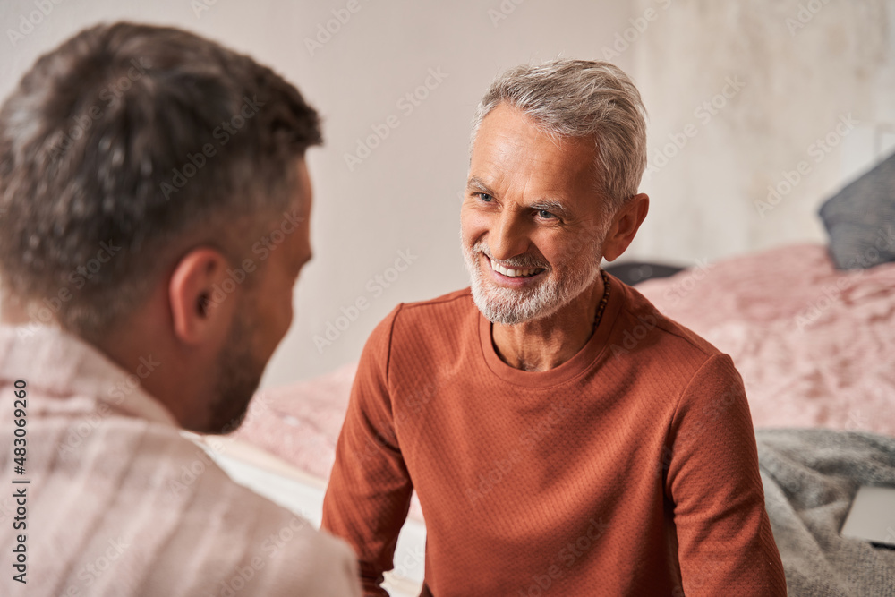 Man remembering something funny with adult son while sitting with each other at home and laughing Man remembering something funny with adult son while sitting with each other at home and laughing